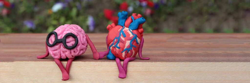 Plasticine human brain and heart sitting on a bench in park and holding hands.